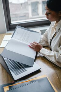 Black woman at desk reading over white paper hard copy in front of computer.